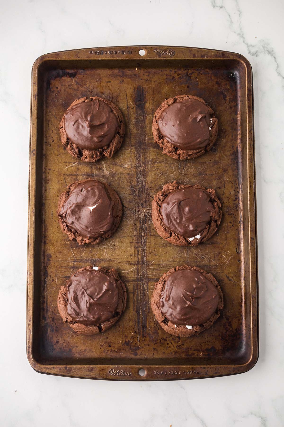 Six large Ding Dong Cookies with frosting on a baking sheet against a white marble background.