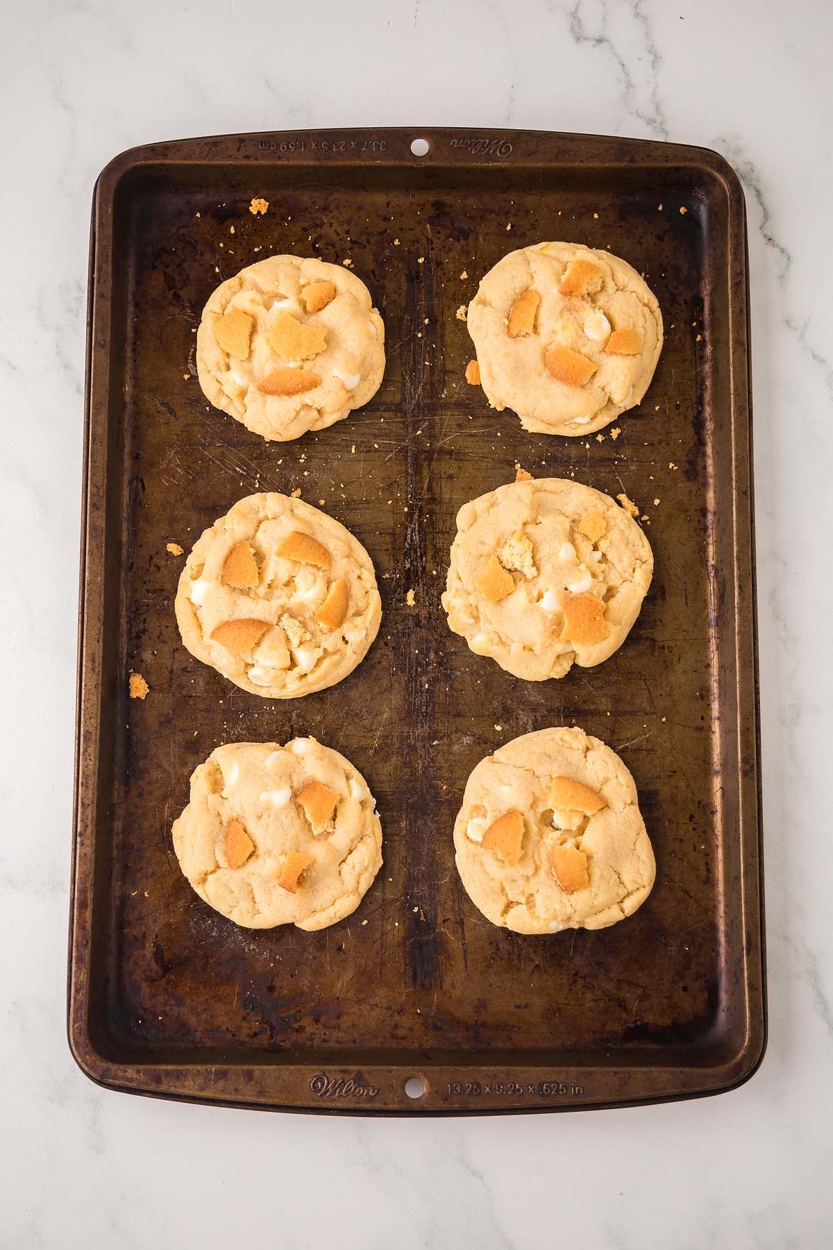 Six Banana Pudding Cookies with white chocolate chips rest on a baking sheet, placed on a white marble surface.