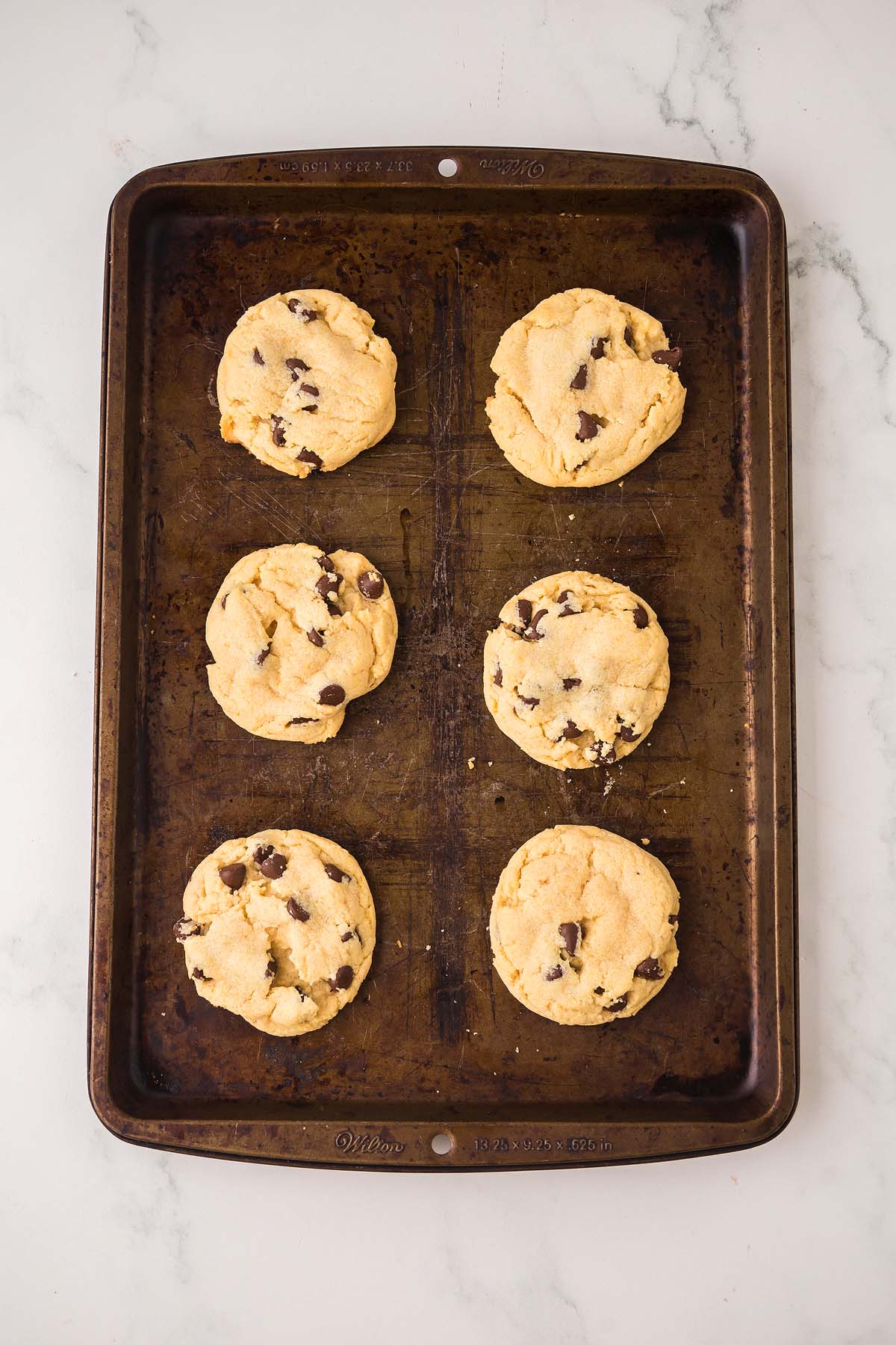 Six Chocolate Chip Pudding Cookies on a dark baking sheet, resting on a white marble surface.