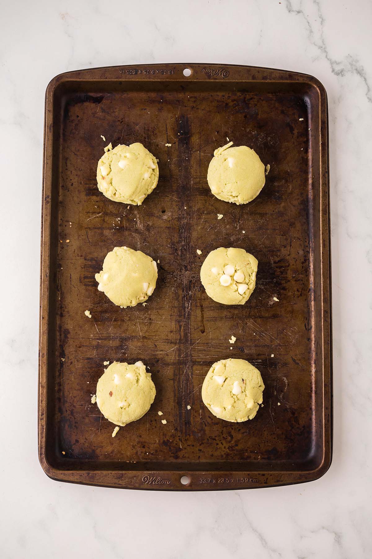 Six unbaked Pistachio Pudding Cookies dotted with white chocolate chips rest on a brown baking sheet, ready to bake.