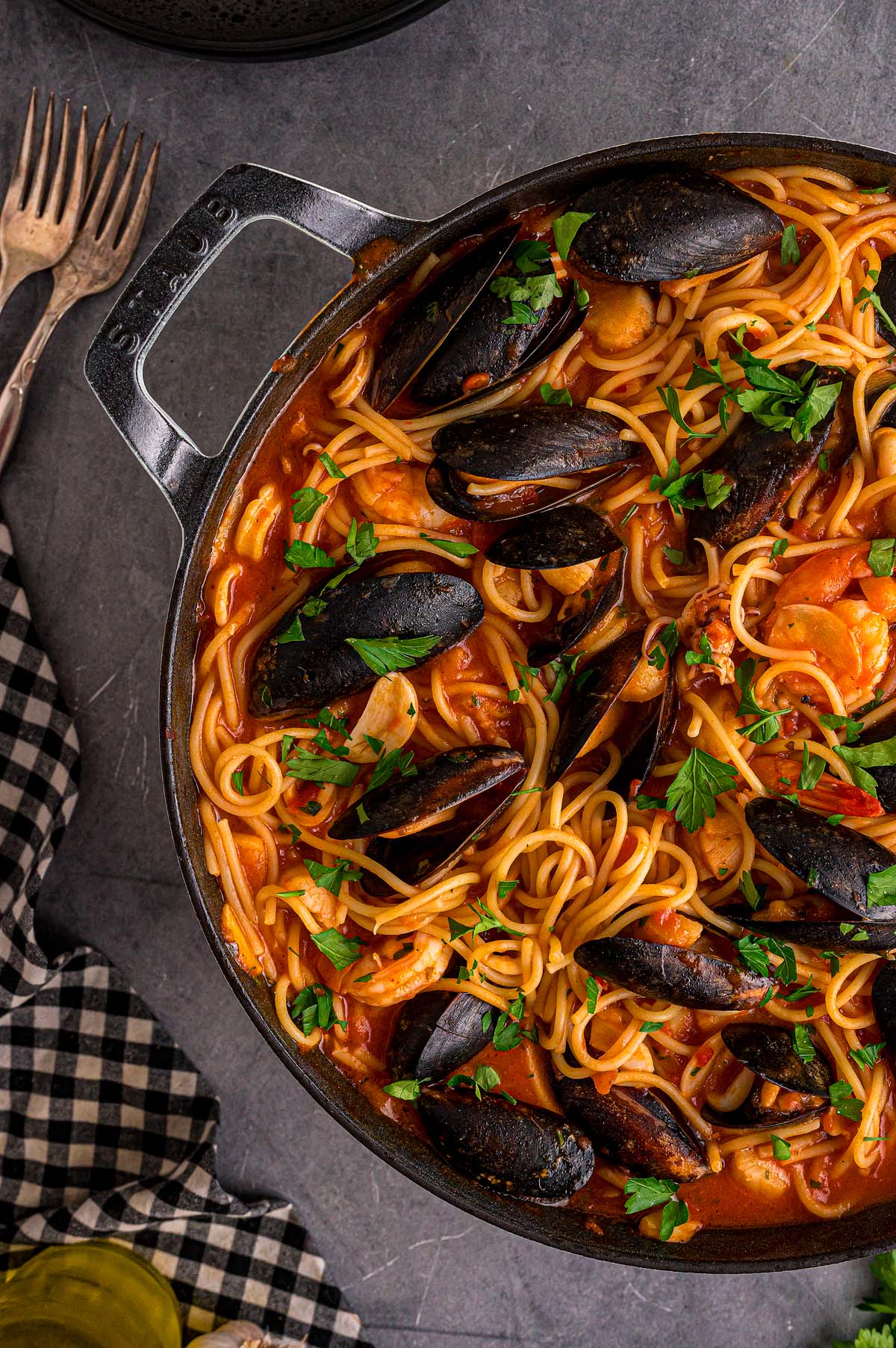 A pot of Frutti di Mare spaghetti with mussels and parsley in tomato sauce, served beside forks and a checked napkin.