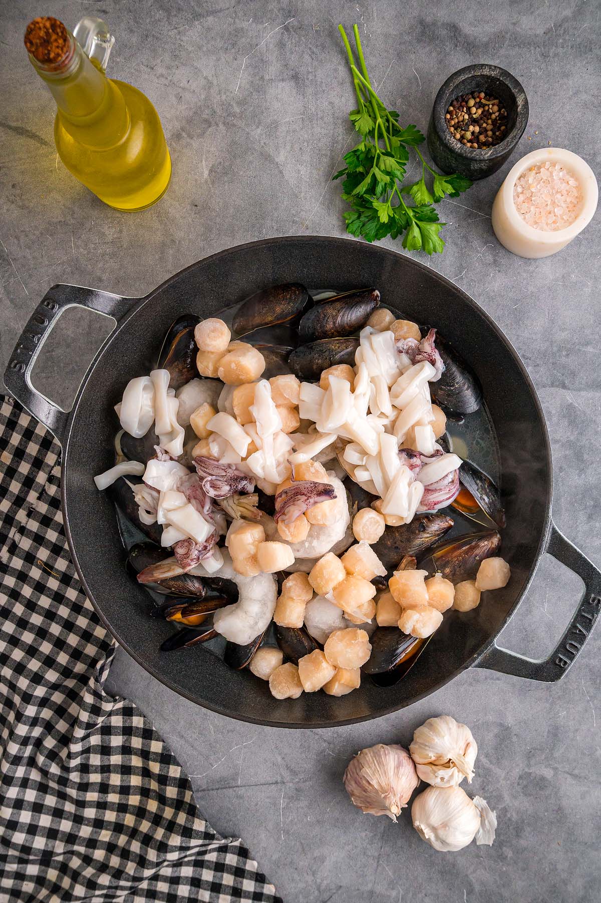 A pan with mixed raw Frutti di Mare seafood, surrounded by garlic, herbs, oil, and seasonings on a gray countertop.