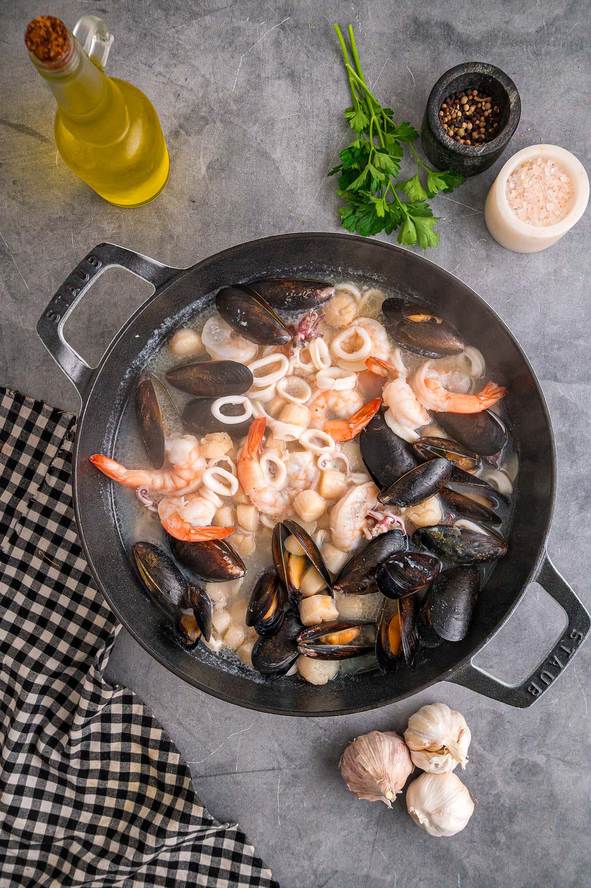A pan of Frutti di Mare, featuring shrimp, mussels, and squid, cooks on a gray surface with fresh herbs and condiments nearby.