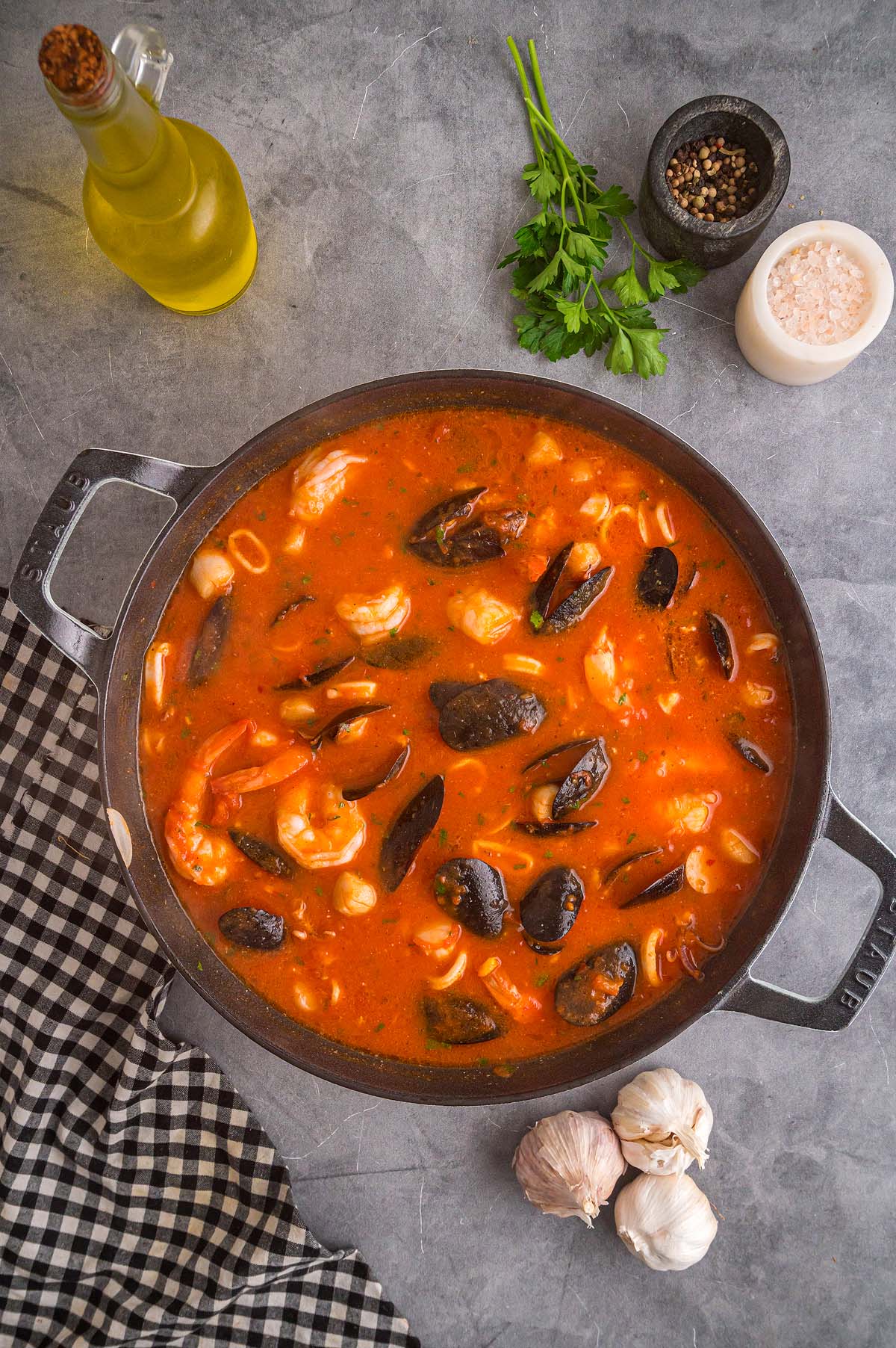 A pot of Frutti di Mare seafood stew with mussels, shrimp, and squid in tomato broth, surrounded by fresh herbs and seasonings.