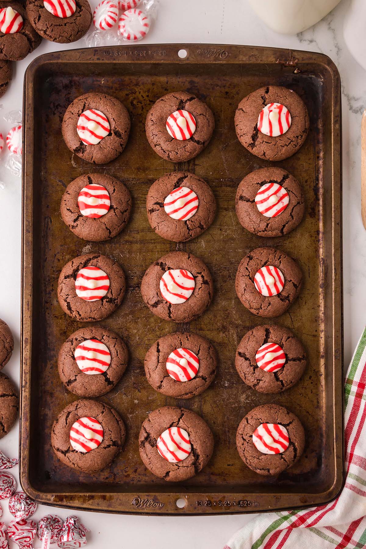 A baking sheet of Peppermint Kiss Cookies—chocolate cookies topped with red and white striped peppermint candies, fresh from the oven.