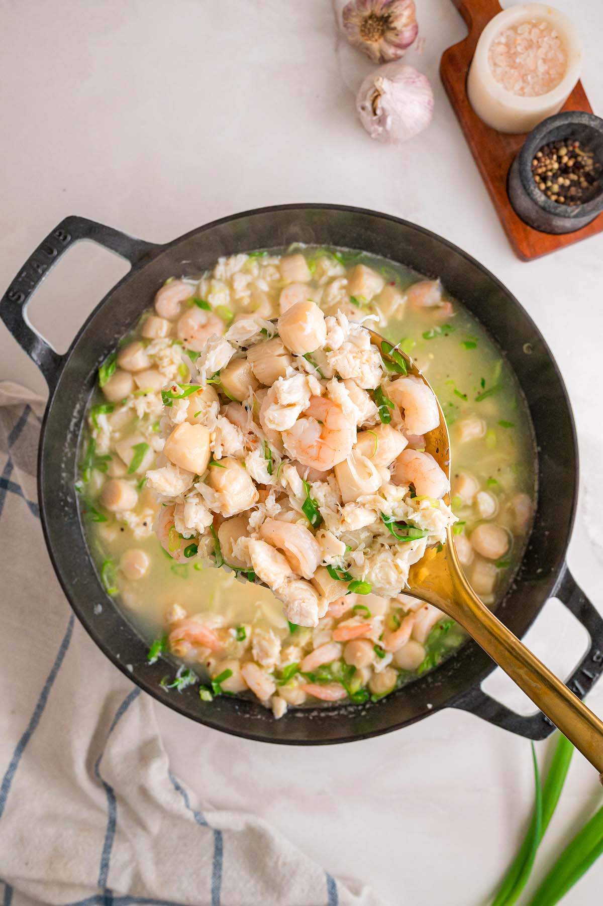 A golden spoon lifts shrimp and seafood from a black pot, surrounded by seasonings on a white surface.