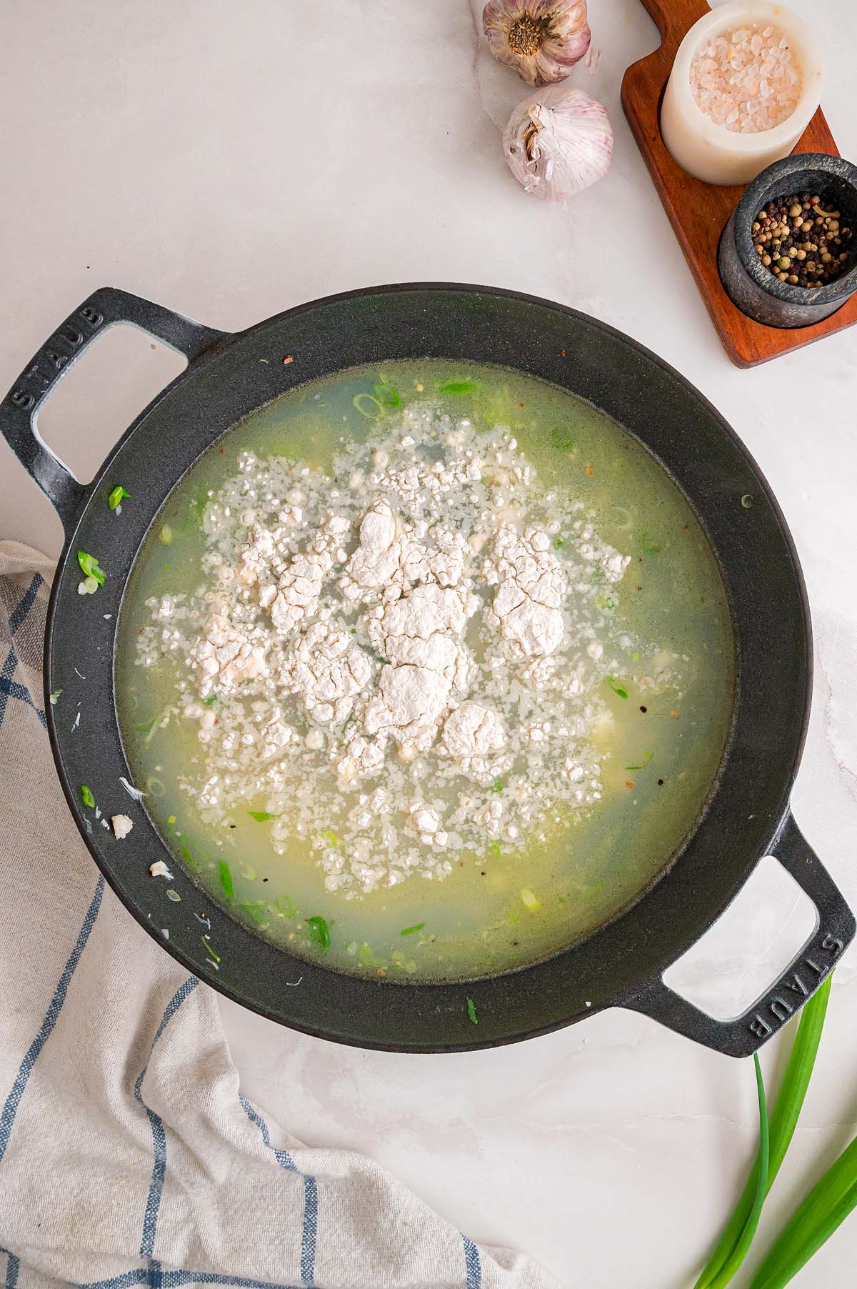 A pot of soup with flour being added, surrounded by garlic, salt, pepper, and a striped towel on the counter.
