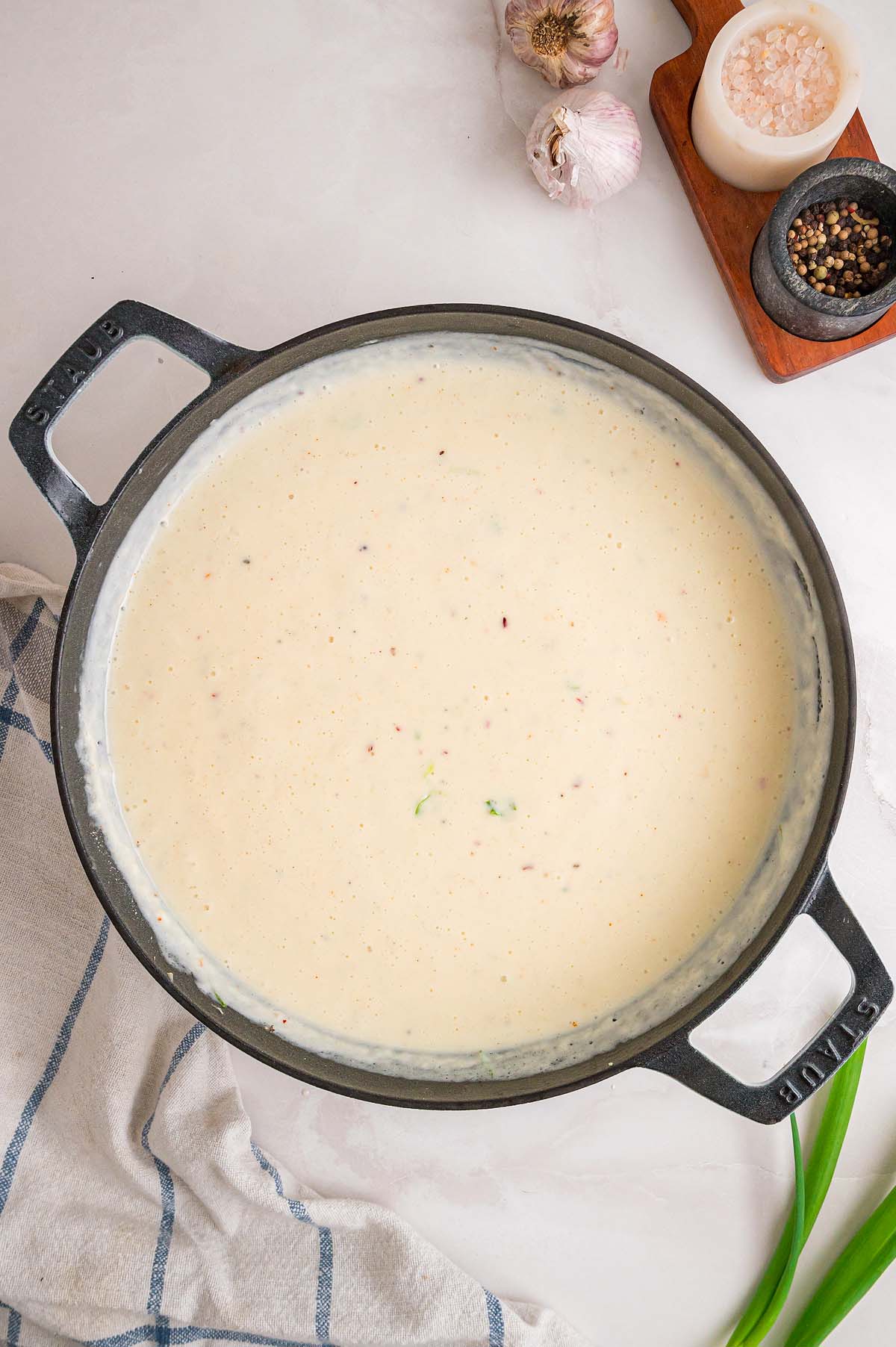 Creamy white sauce with herbs in a black pot, surrounded by salt, pepper, garlic, and a striped towel.