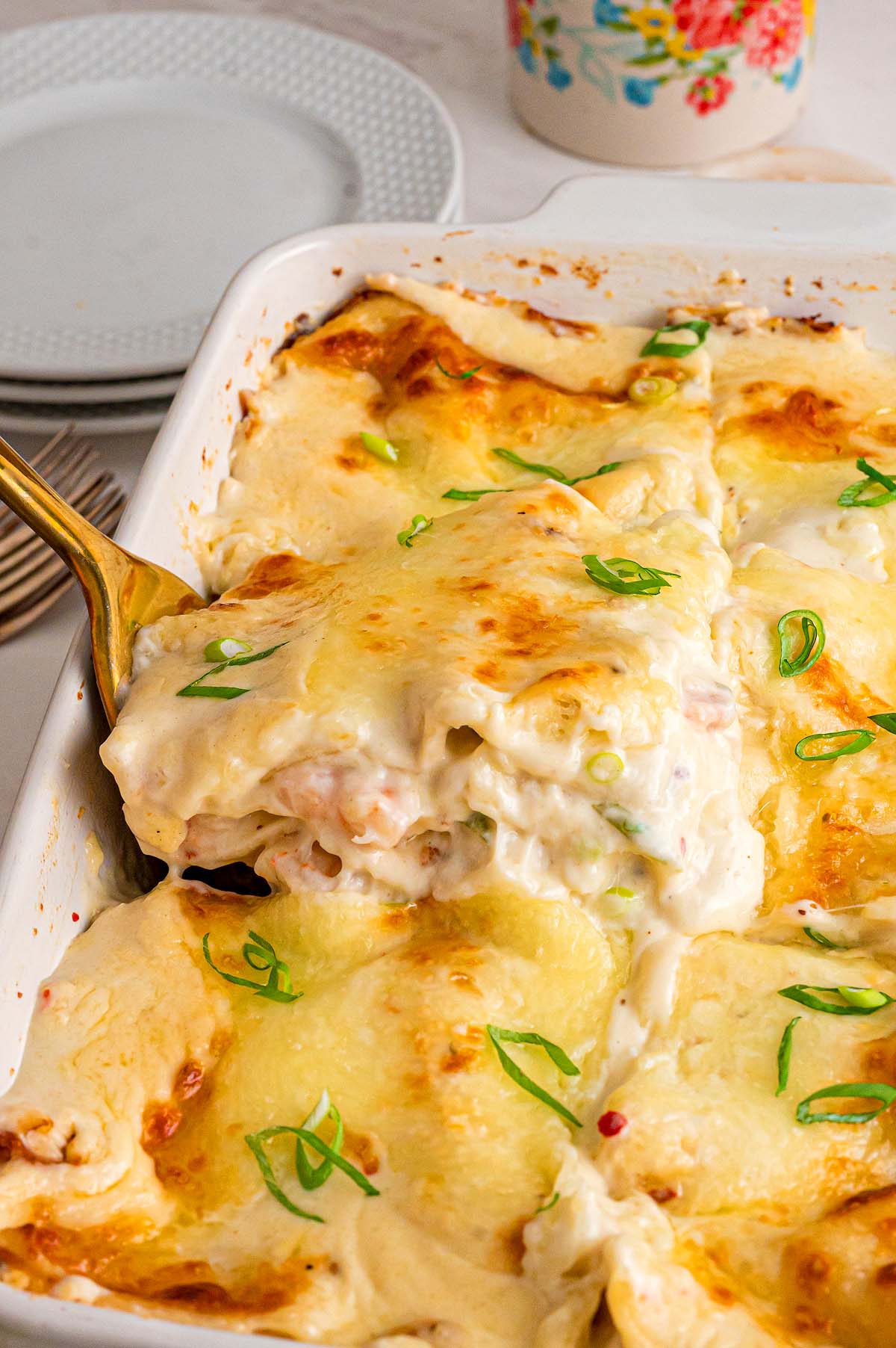 Golden, cheesy seafood lasagna topped with green onions, served from a white baking dish with plates in the background.