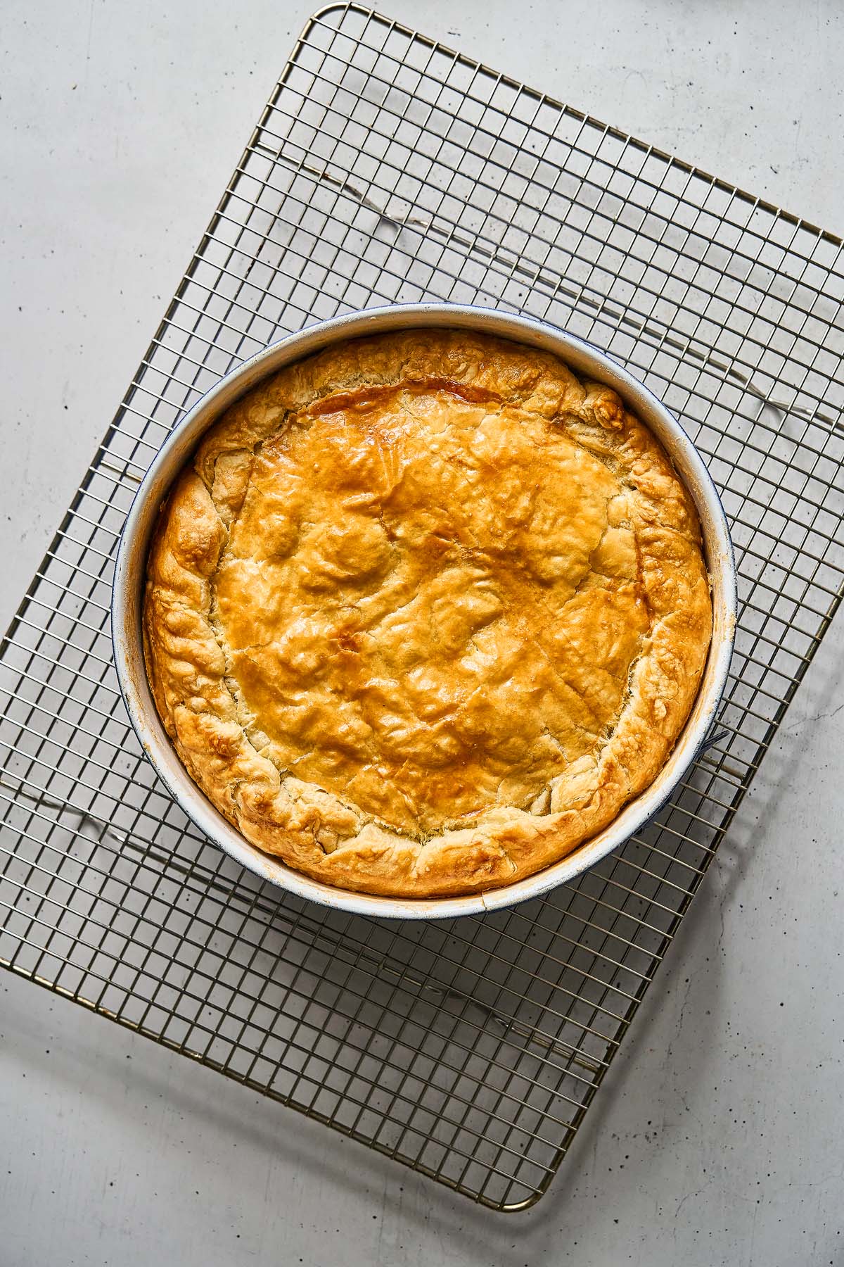 A golden-brown Torta Pasqualina in a round pan rests on a metal cooling rack over a light-colored surface.