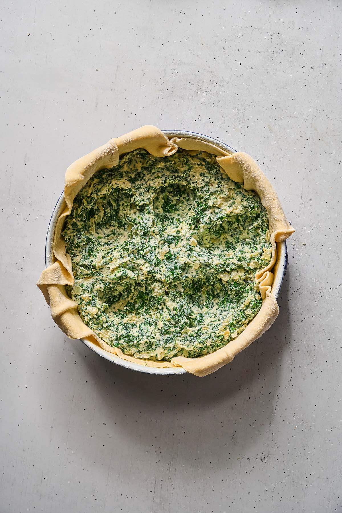 Unbaked Torta Pasqualina in a round pan, with flaky pastry edges folded over a rich spinach and ricotta filling.
