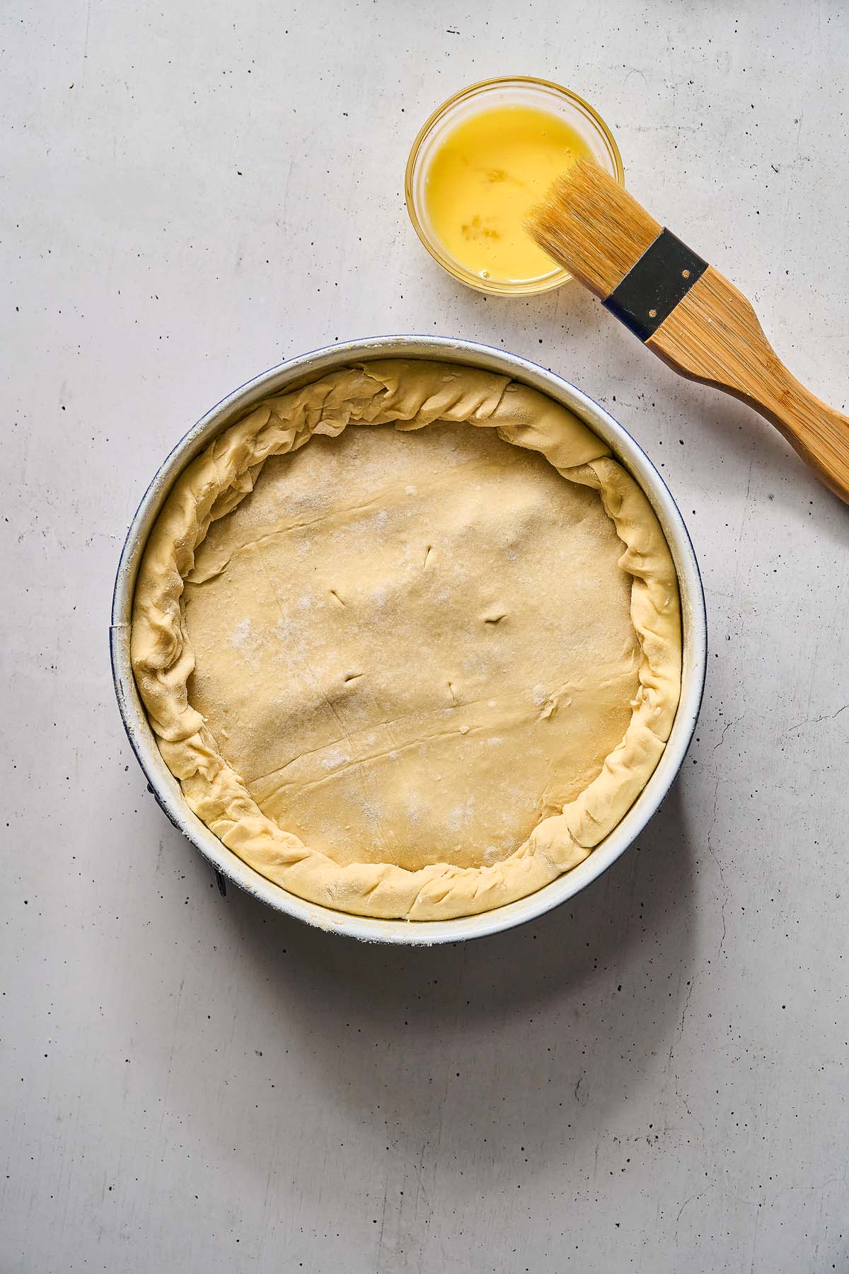 Unbaked Torta Pasqualina with crimped edges in a round pan beside a bowl of egg wash and a pastry brush.