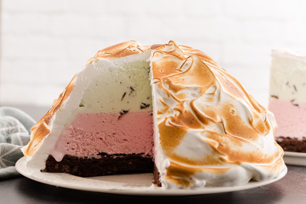 A sliced Baked Alaska cake with layers of ice cream and perfectly browned meringue sits elegantly on a white plate.