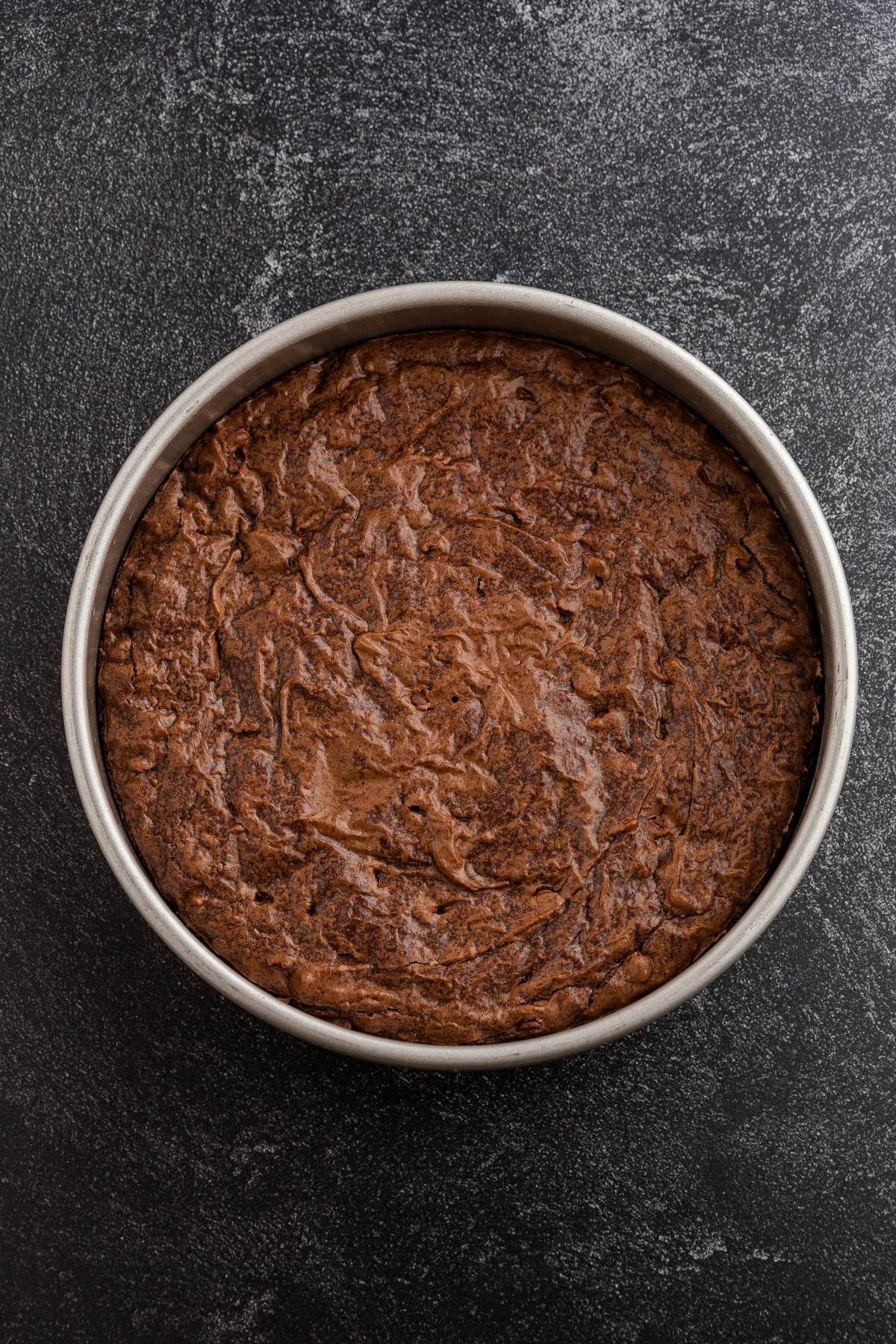 A round chocolate cake in a metal baking pan on a dark textured surface.