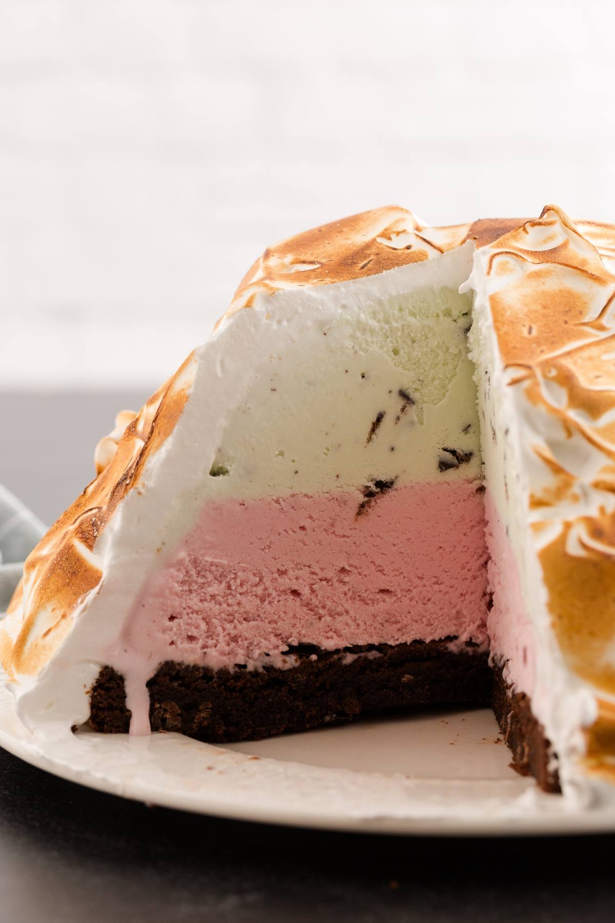 A sliced Baked Alaska showing layers of green and pink ice cream on a chocolate base, topped with toasted meringue.