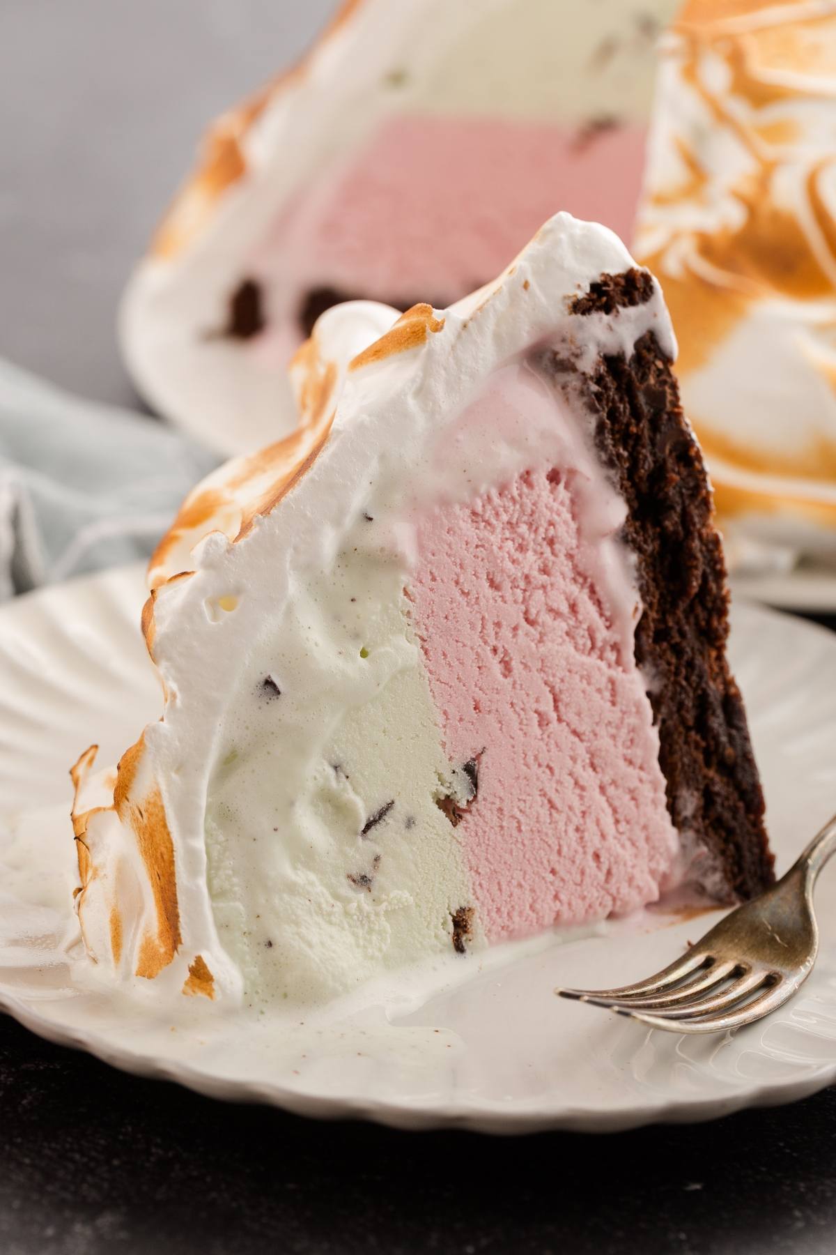 A slice of baked Alaska with chocolate cake, pink and green ice cream, and toasted meringue on a white plate.