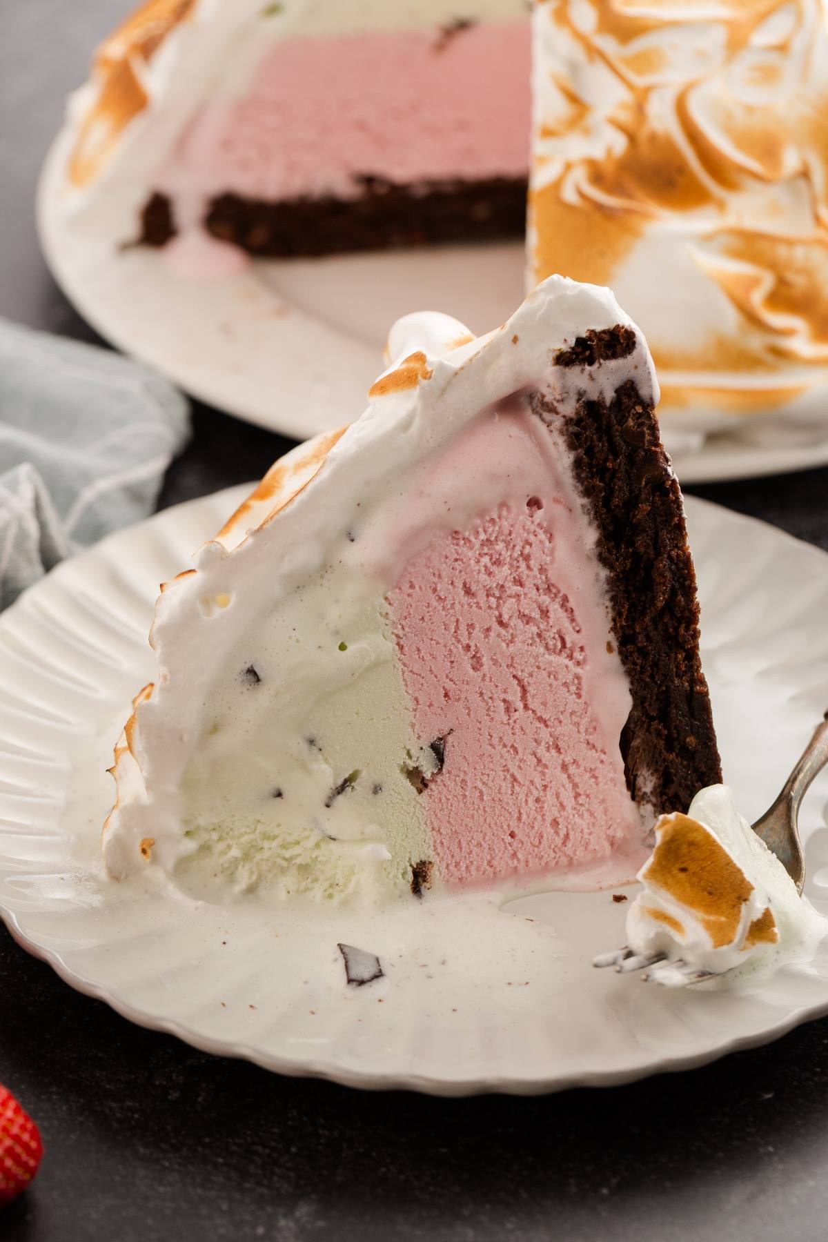 Slice of baked Alaska with layers of chocolate cake, strawberry and mint ice cream, topped with toasted meringue.