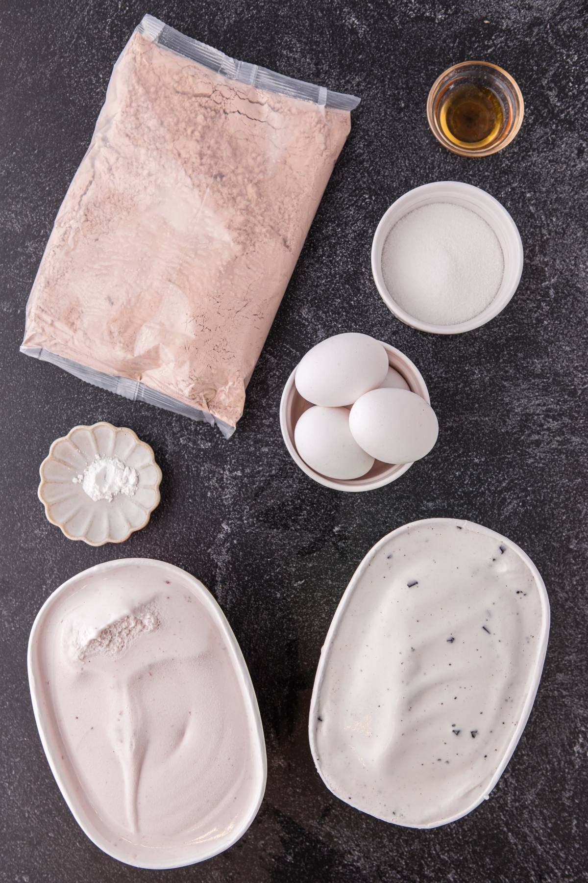 Two tubs of ice cream, eggs, sugar, vanilla, baking powder, and a bag of cake mix on a dark surface.