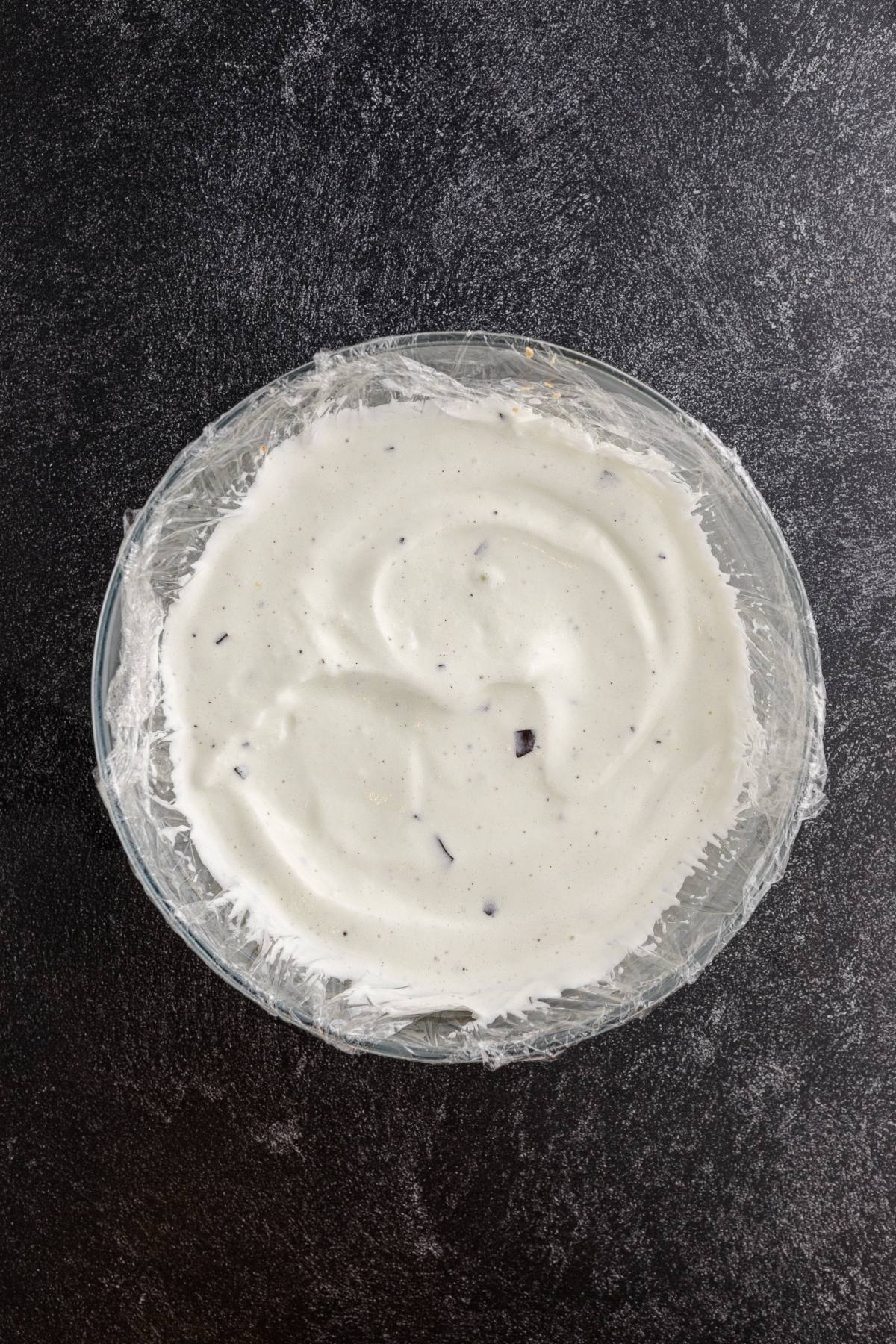 A glass bowl filled with white ice cream mixture, covered with plastic wrap, on a dark surface.