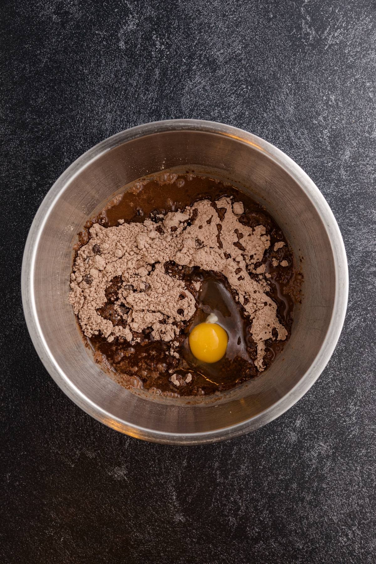 A metal bowl with brownie mix, an egg, water, and oil on a dark countertop.