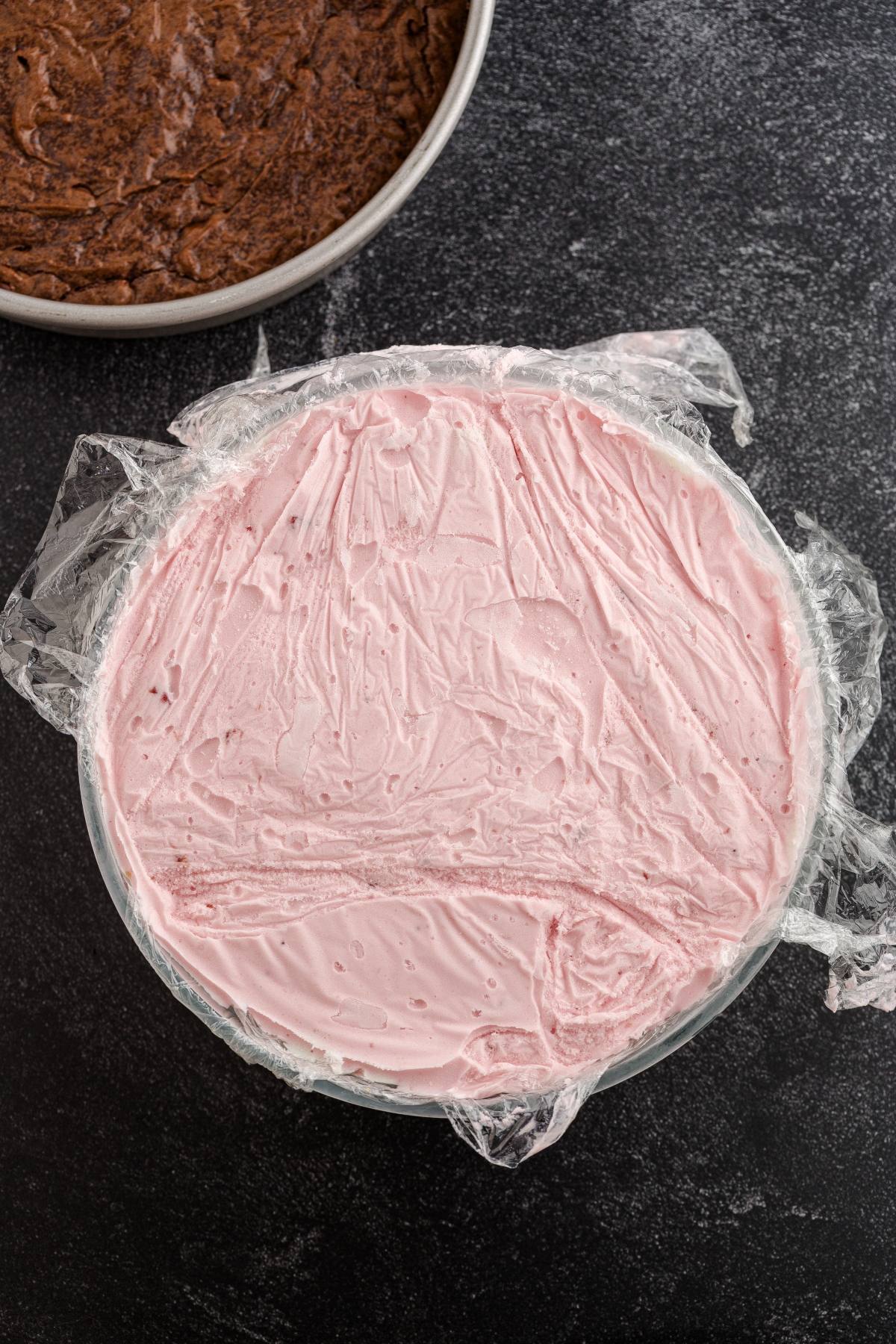 A bowl of pink ice cream covered with plastic wrap, next to a pan of chocolate batter on a dark surface.