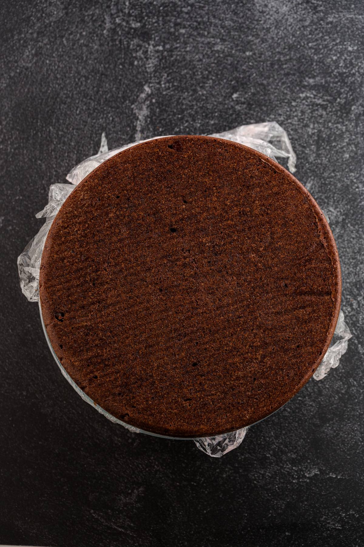 A round chocolate cake on a dark surface, wrapped with plastic at the edges.