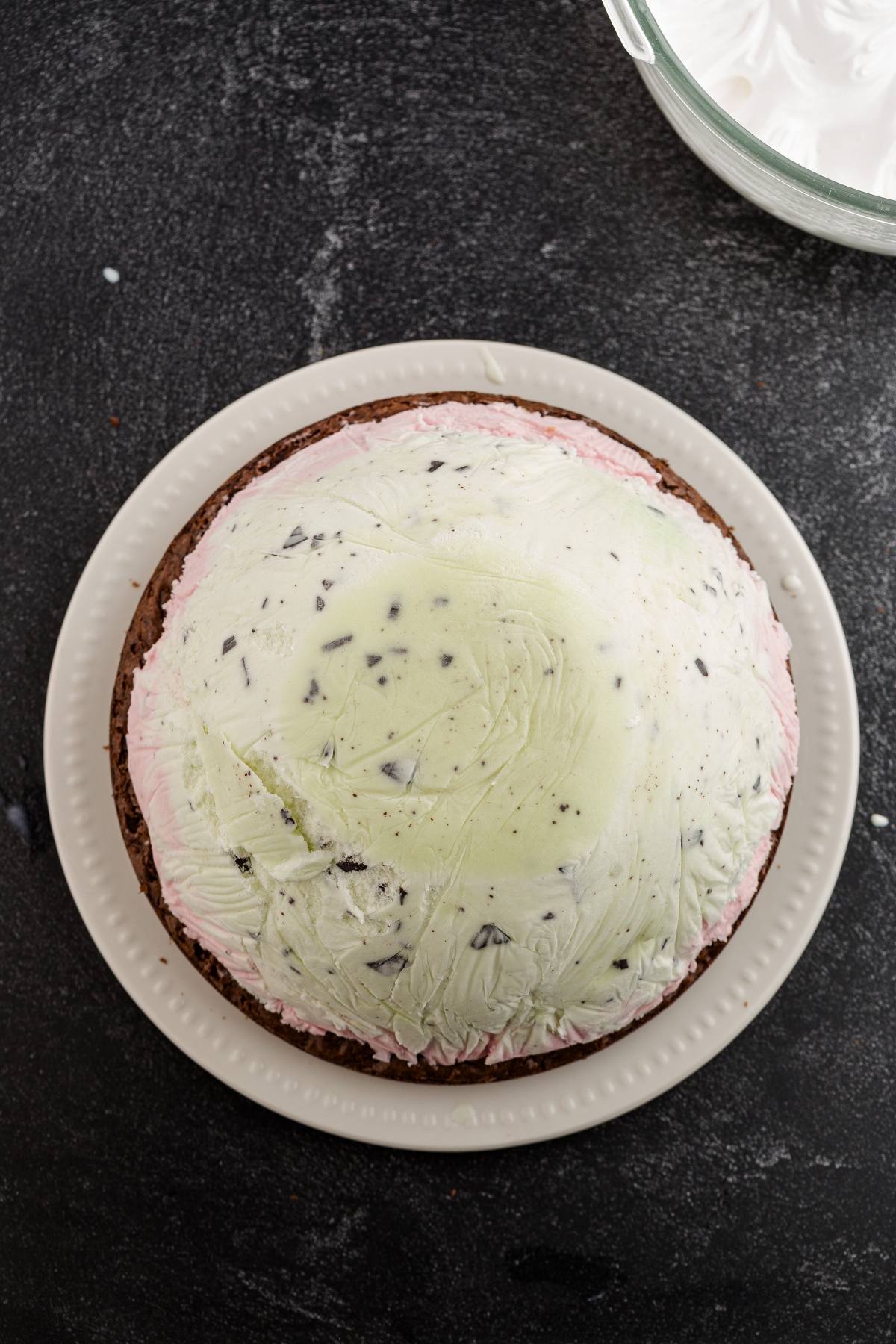 A round ice cream cake with chocolate, pink, and green layers on a white plate, seen from above.