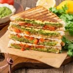 Stacked Bombay sandwiches with grilled vegetables, green chutney, tomatoes, and fresh cilantro on a wooden board.