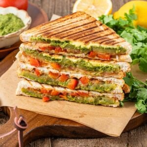 Stacked Bombay sandwiches with grilled vegetables, green chutney, tomatoes, and fresh cilantro on a wooden board.
