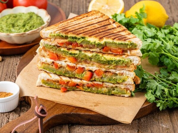 Stacked Bombay sandwiches with grilled vegetables, green chutney, tomatoes, and fresh cilantro on a wooden board.