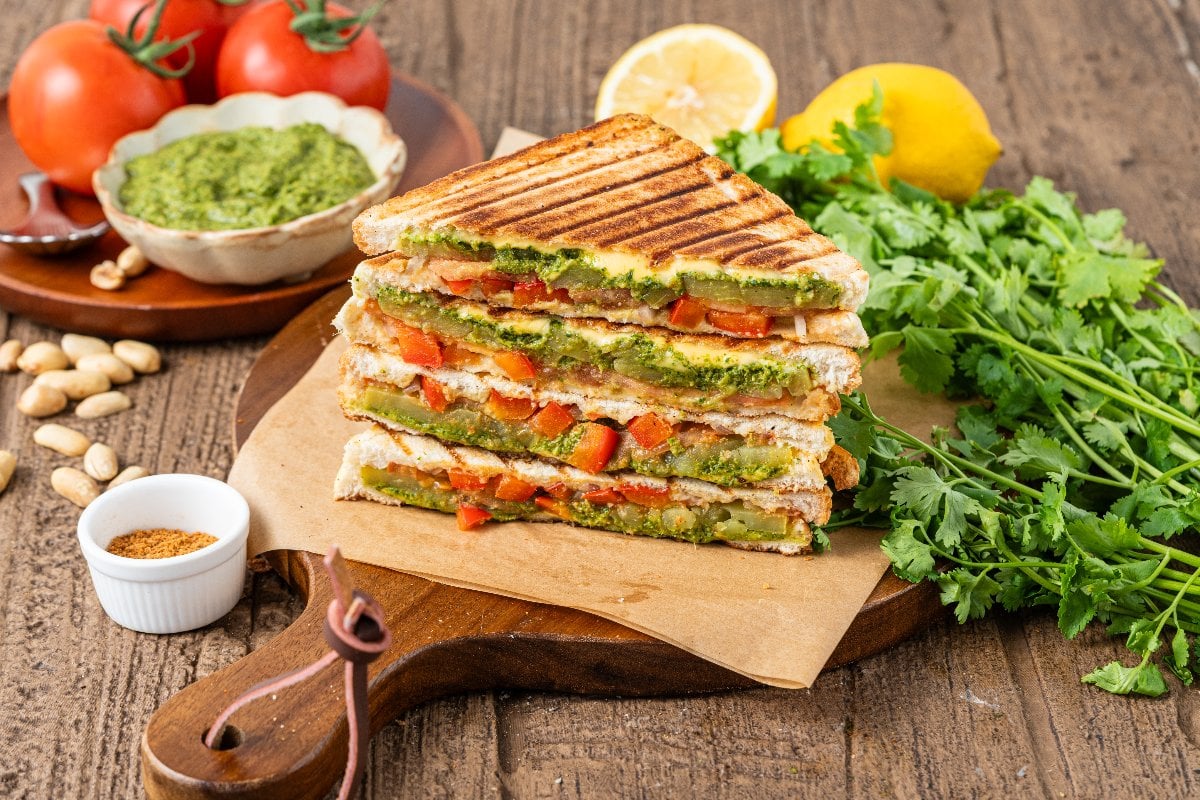 Grilled Bombay veggie sandwiches stacked on a board, with fresh herbs, tomatoes, and a bowl of green pesto nearby.
