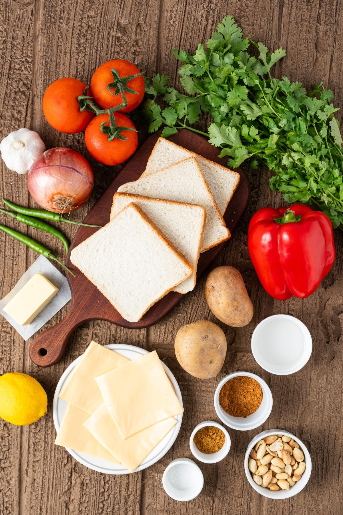 Sandwich ingredients: sliced bread, cheese, veggies, spices, herbs, butter, peanuts, and lemon on a wooden table.