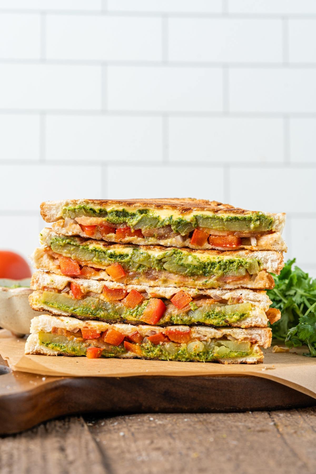 Stacked veggie bombay sandwiches with grilled bread, filled with tomato, greens, and spread, on a wooden board.