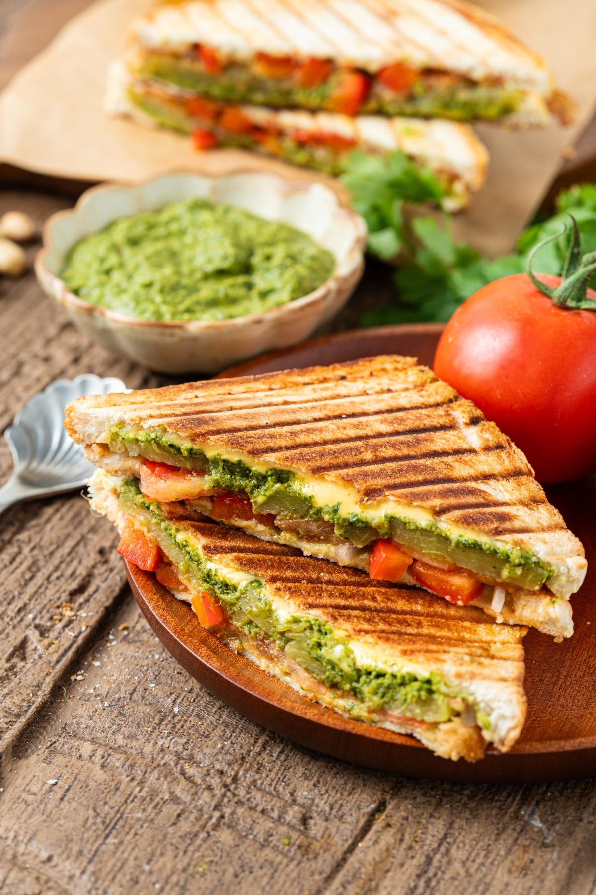 Grilled vegetable bombay sandwich with pesto, sliced tomatoes, and a bowl of green pesto sauce on a wooden table.