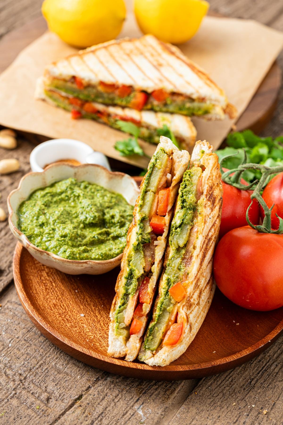 Grilled vegetable bombay sandwich with green pesto dip, tomatoes, and lemons on a wooden plate and table.