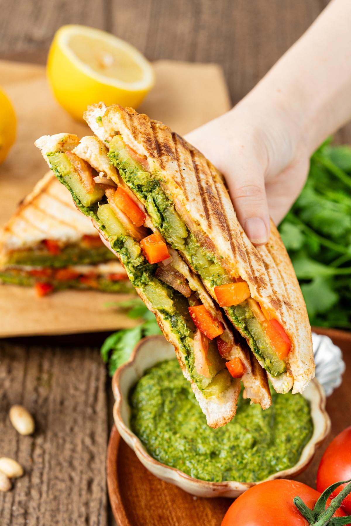 A hand holds a grilled bombay vegetable sandwich with pesto sauce, with fresh veggies and lemon in the background.