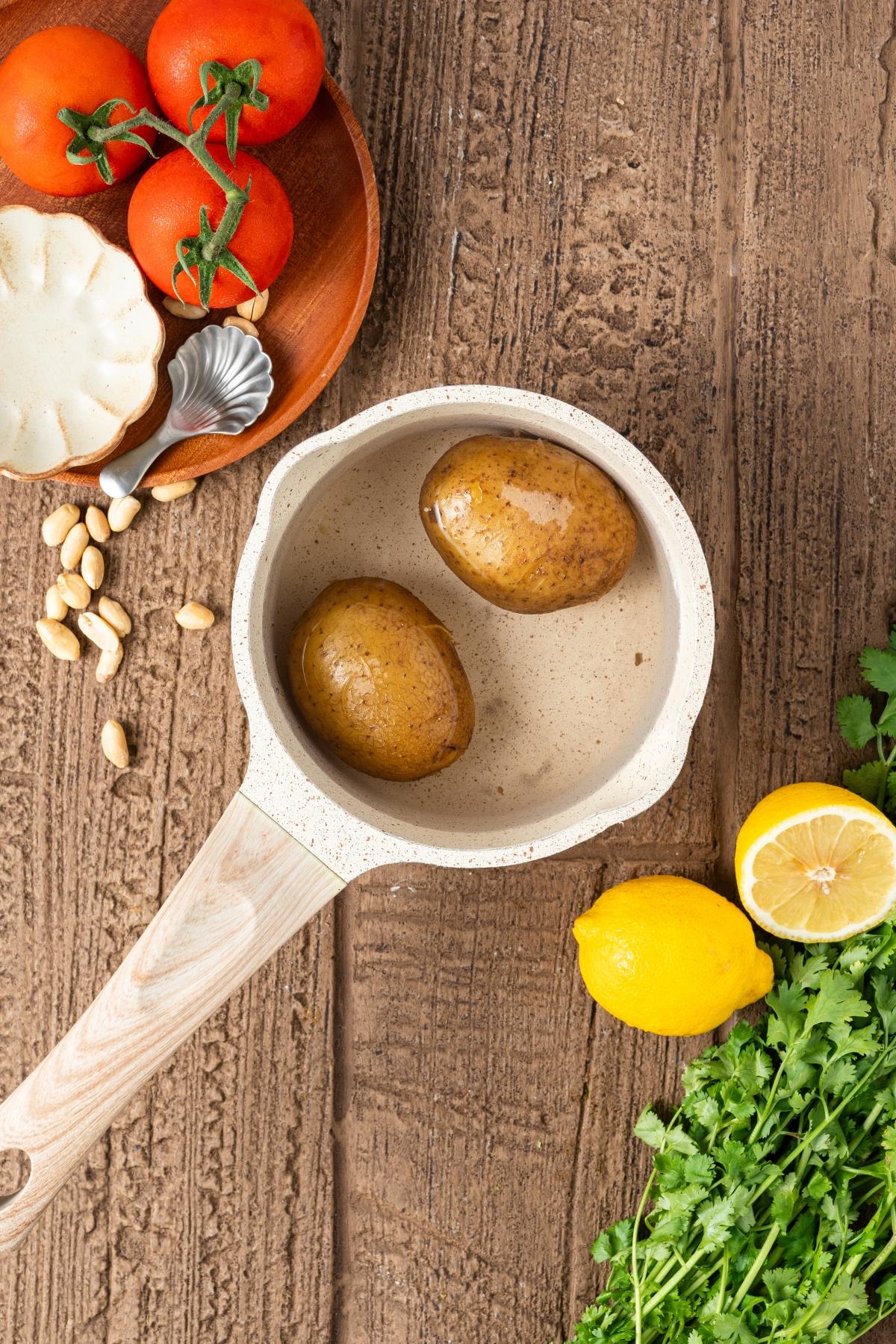 A saucepan with two potatoes, surrounded by tomatoes, garlic, lemon, and fresh cilantro on a wooden surface.