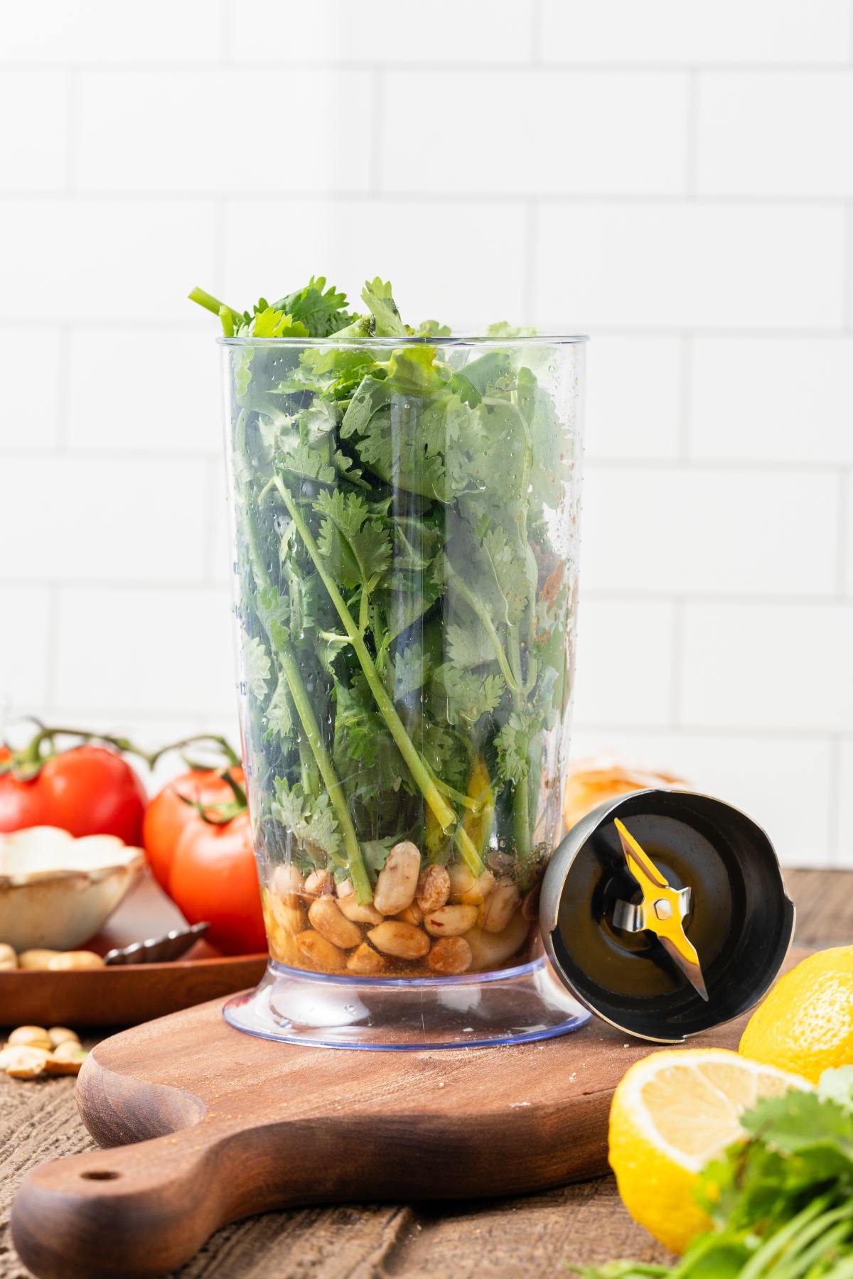 A blender cup filled with cilantro and nuts sits on a board, with tomatoes and lemons nearby.