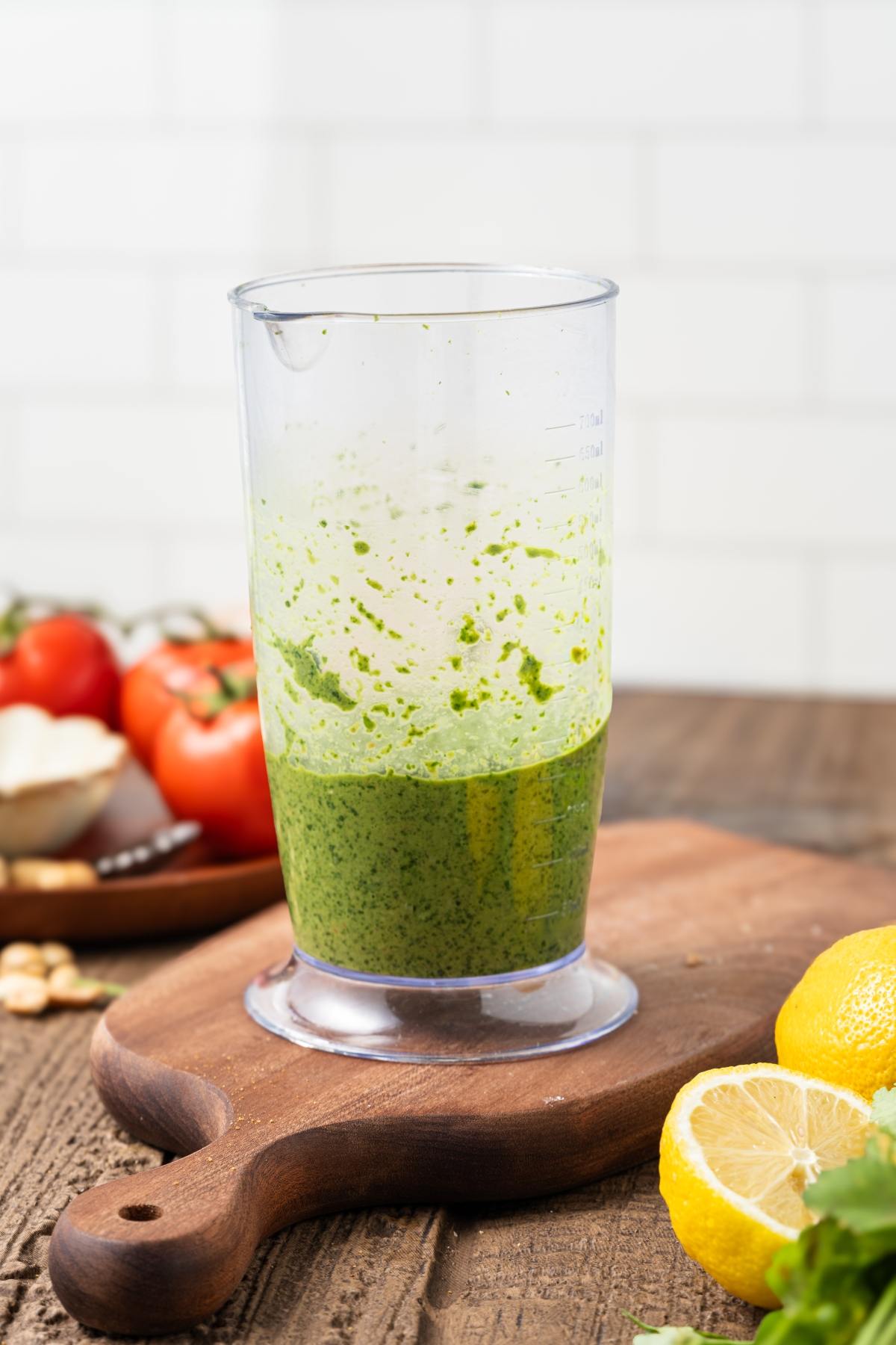 Clear blender cup with green pesto on a wooden board, lemon halves and fresh veggies in the background.