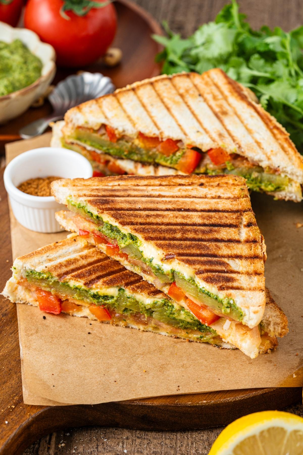Grilled vegetable bombay sandwiches with pesto, tomatoes, and cheese, cut in halves and served on parchment paper.