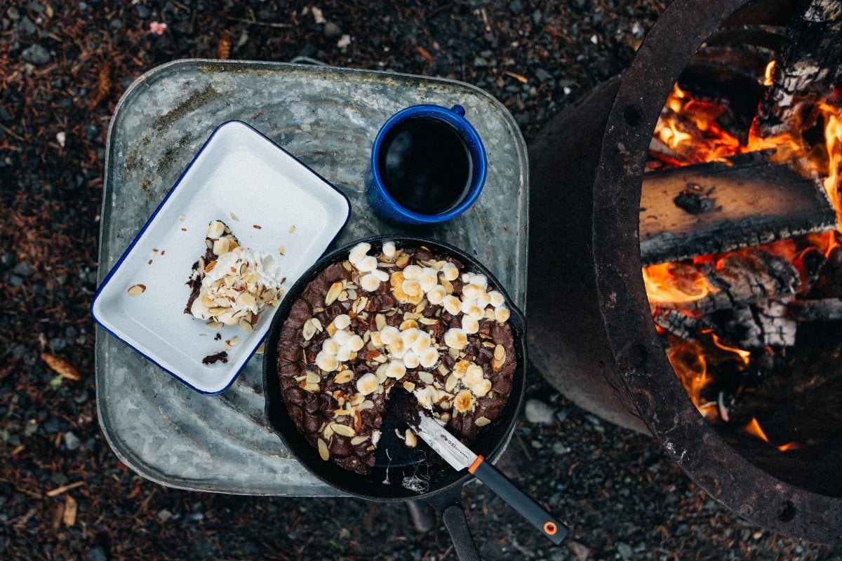 A Campfire Cast Iron Caramel Brownie dessert topped with marshmallows and nuts sits beside coffee by a campfire, with a slice served on a plate.