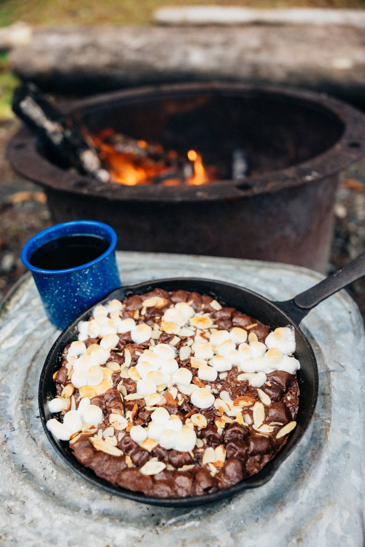 A campfire cast iron caramel brownies dessert topped with marshmallows beside a blue mug, near a campfire.