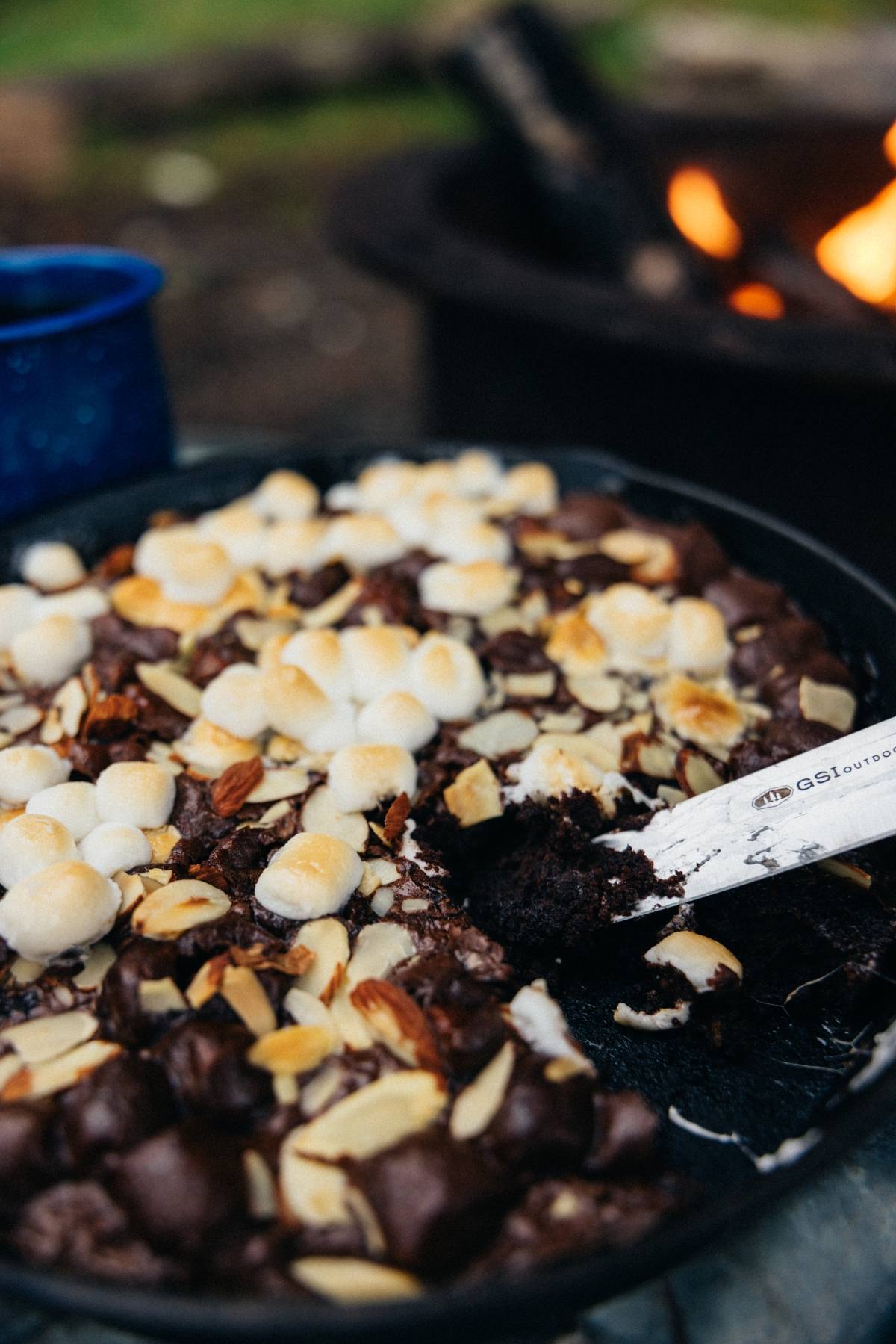 A campfire cast iron caramel brownies dessert with toasted marshmallows and almonds beside a campfire, a spoon serving a portion.