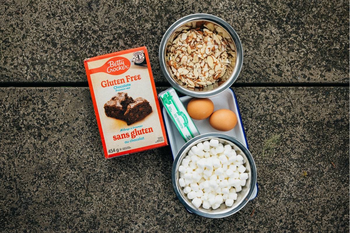 Brownie mix, sliced almonds, eggs, butter, and marshmallows arranged on a speckled concrete surface.