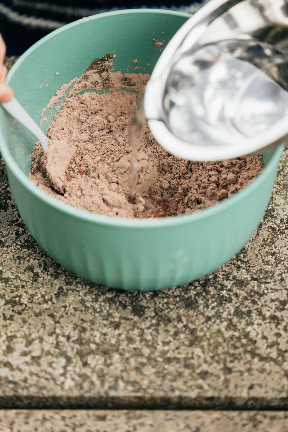 A person pours water into a green bowl of cocoa powder using a metal pitcher on a stone surface.