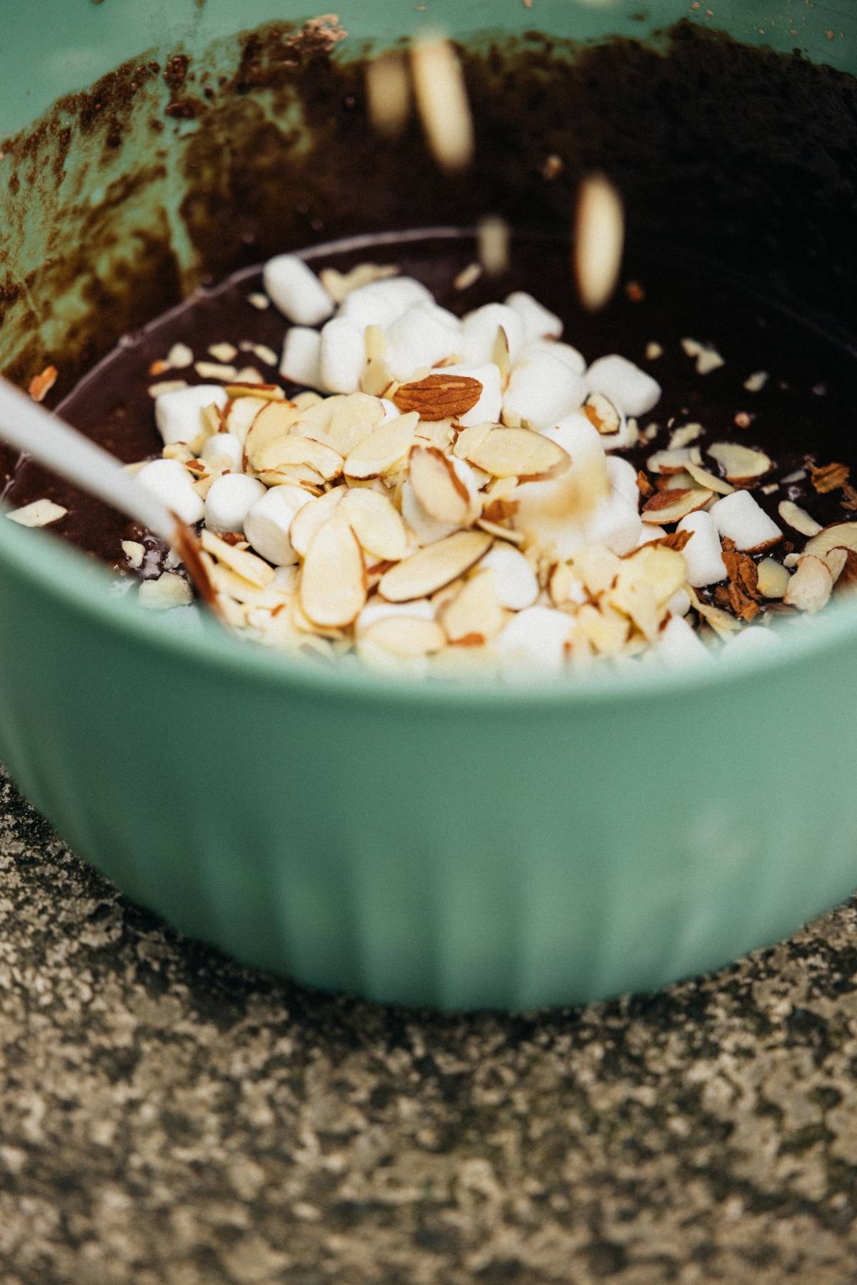 A green bowl with chocolate batter, mini marshmallows, and sliced almonds being mixed.