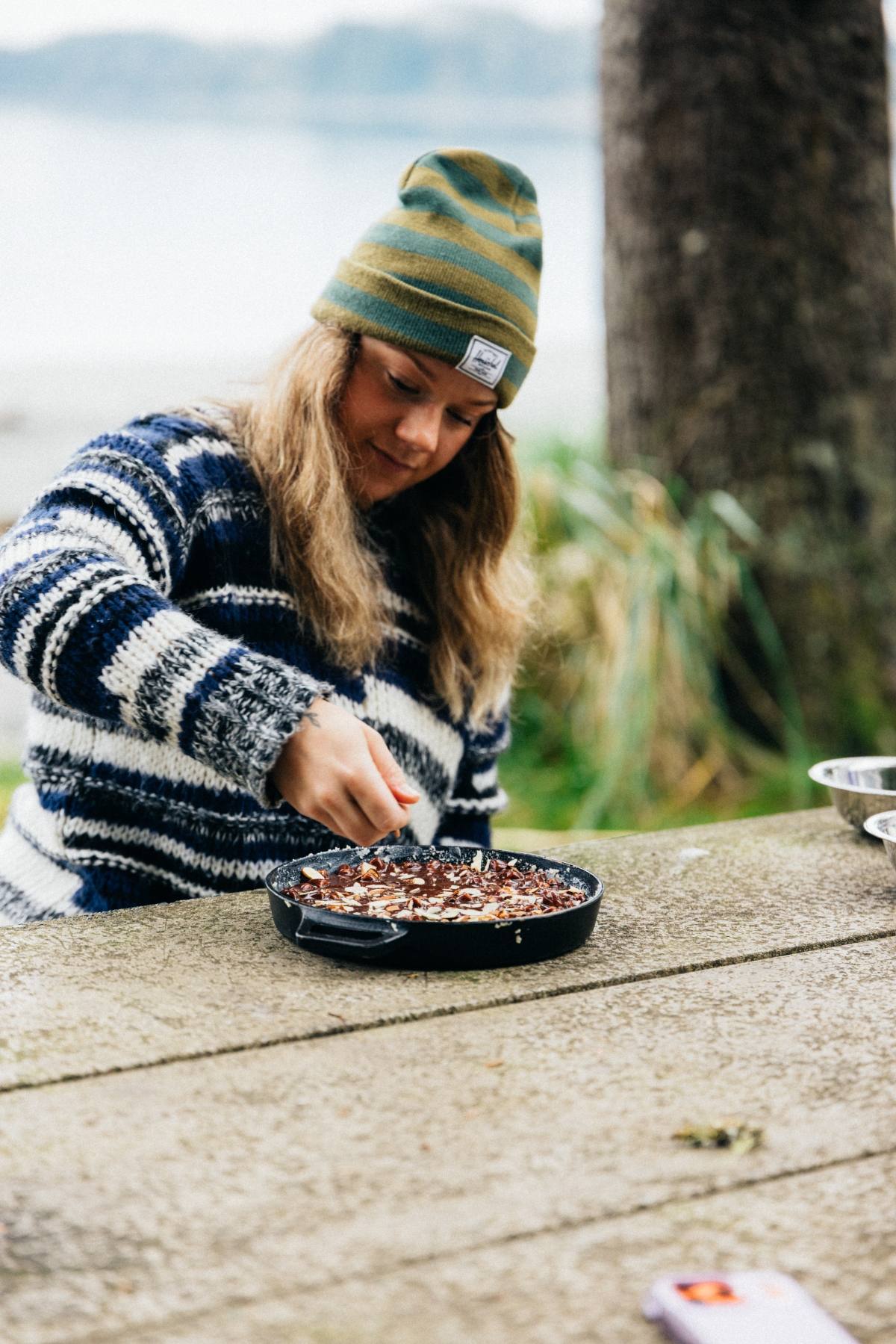 A person in a striped sweater and beanie sits outdoors, stirring food in a black pan on a wooden table.