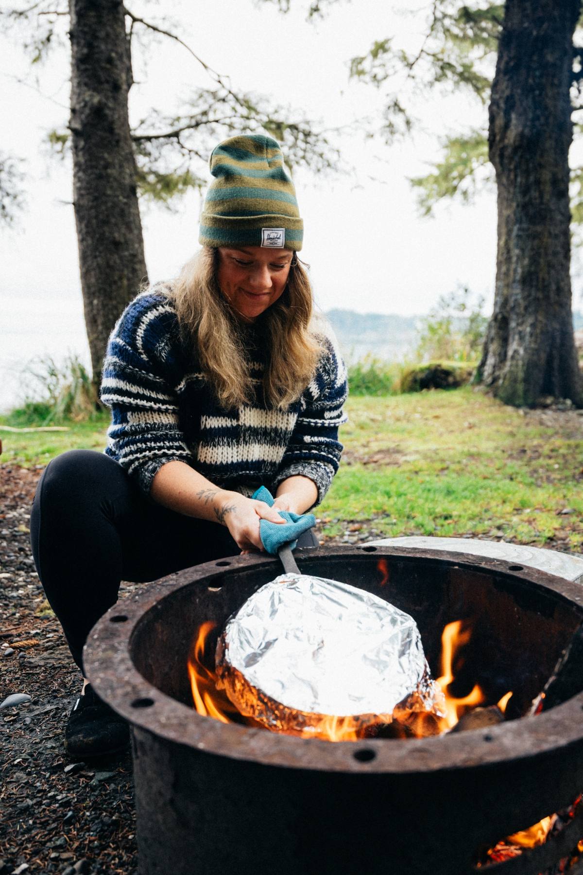 Person in a striped sweater and beanie tending to foil-wrapped food over a campfire outdoors.