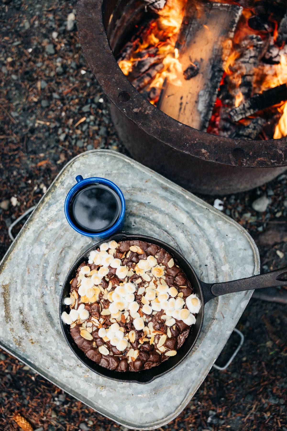 A campfire cast iron caramel brownies with dessert topped with marshmallows and a mug of coffee beside a campfire.