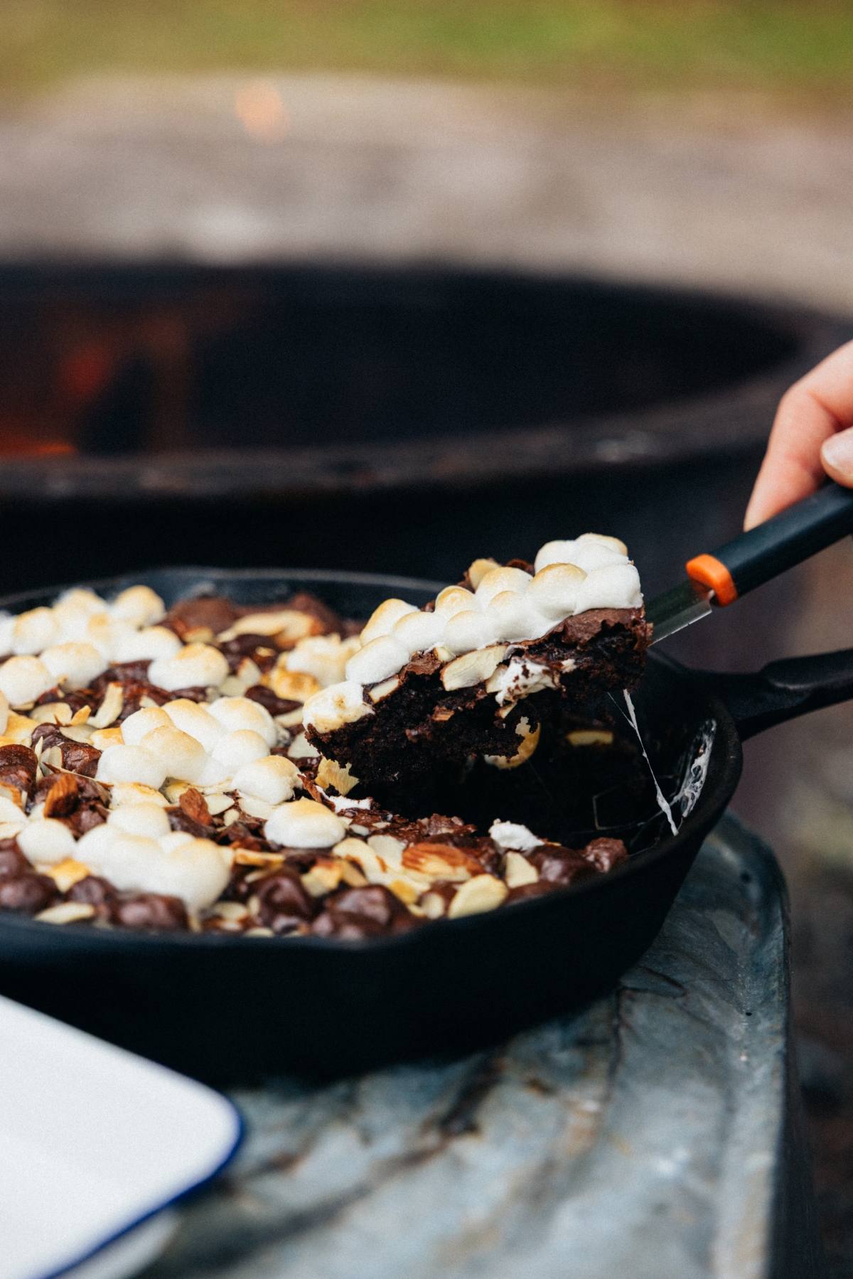 A campfire cast iron caramel baked brownies topped with melted marshmallows, with a spatula lifting a serving.